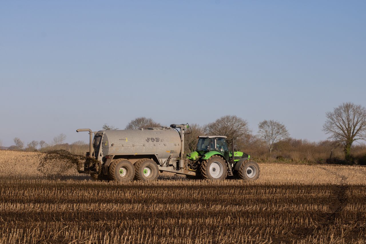 A green tractor spreading fertilizer on a brown, freshly plowed farmland under a clear sky.